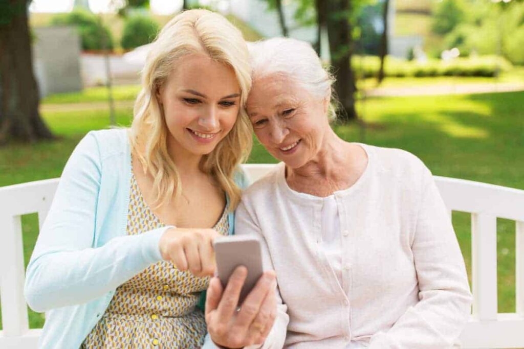 Senior woman sitting outdoors on a bench with her daughter looking at information for Traditions of Mount Vernon on a smartphone