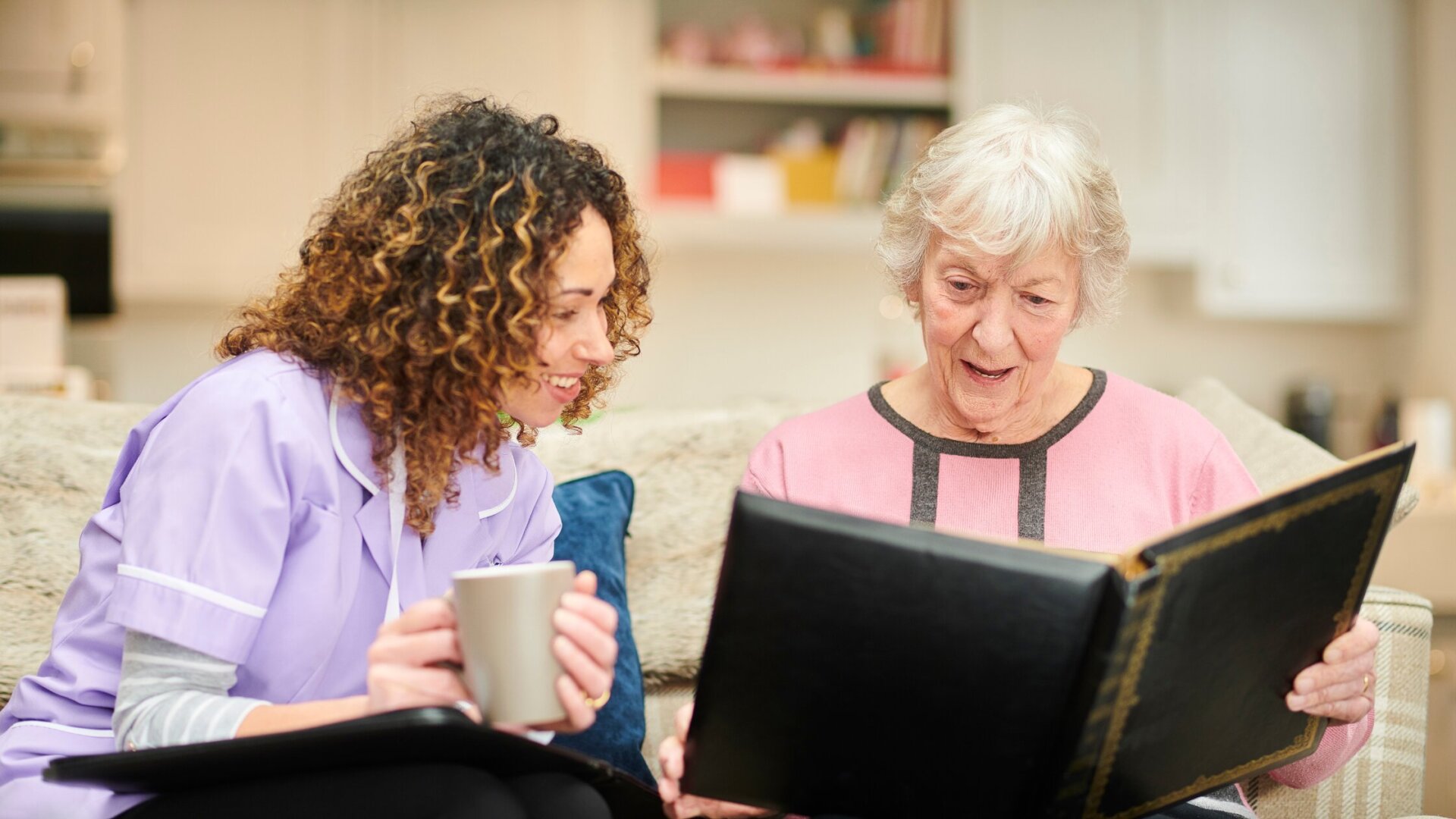 Female memory care resident sitting on a couch in her memory care apartment at Traditions of Mount Vernon with a caregiver