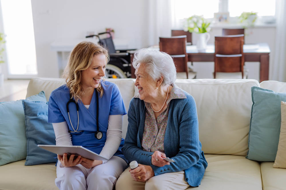 Female nurse sitting on a sofa with a memory care patient, laughing and conversing, at Traditions of Mount Vernon in Mount Vernon, OH.